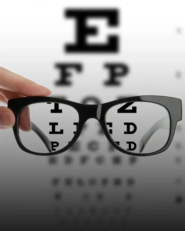 A pair of glasses held up in front of a blurry eye chart. The chart is clear seen through the glasses lenses.