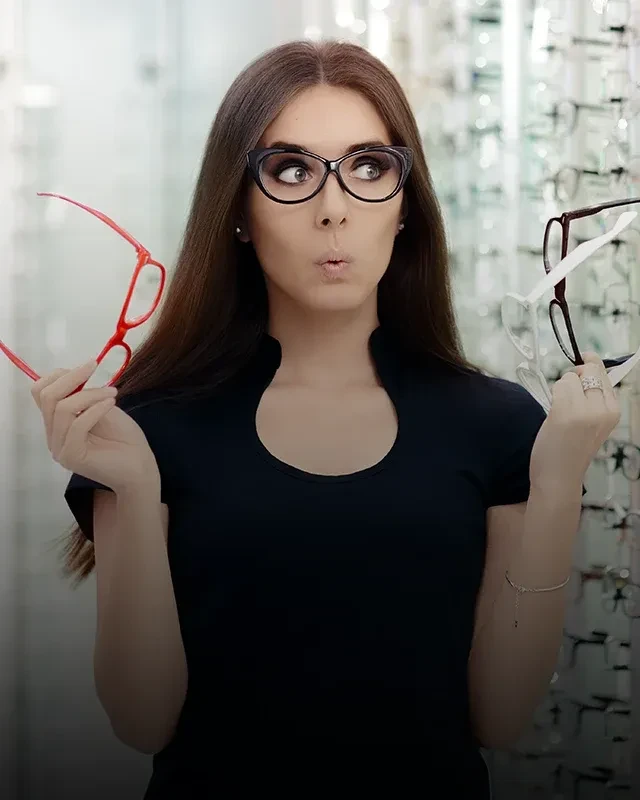 A woman holding multiple pairs of eyeglass frames while looking at a wall-mounted frames display